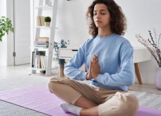 A women with brown curly hair sits cross legged on a yoga mat with her eyes closed in a meditative pose